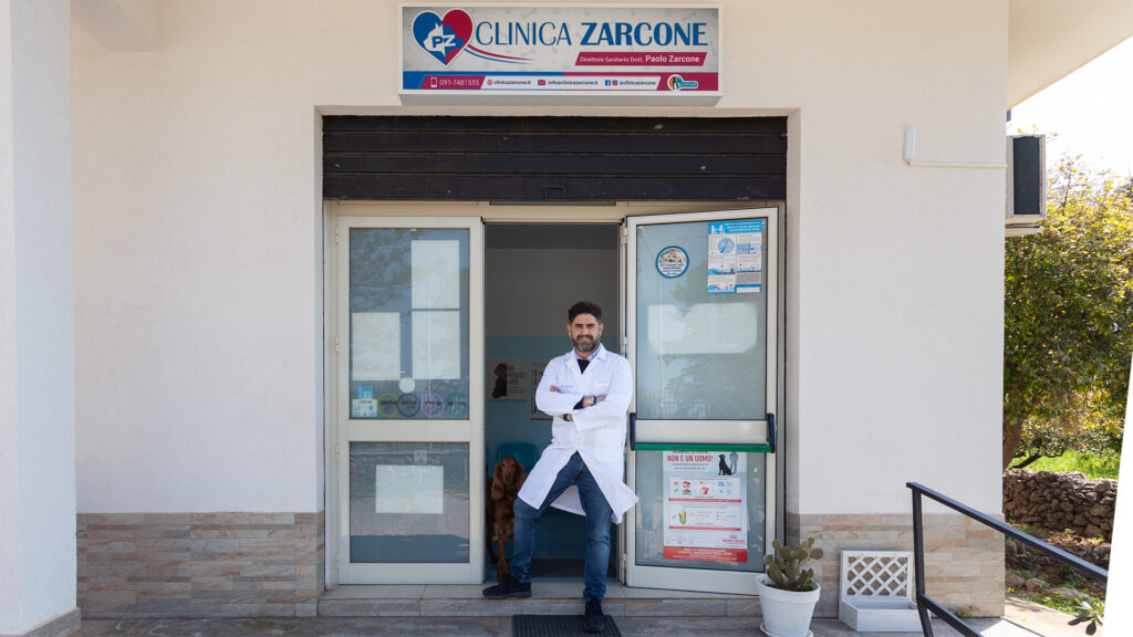 cinisi_05 Un uomo con un camice bianco sta a braccia incrociate all'ingresso della Clinica Zarcone, un ambulatorio medico con porte di vetro e un'insegna sopra l'ingresso. A destra c'è una piccola pianta e delle scale.