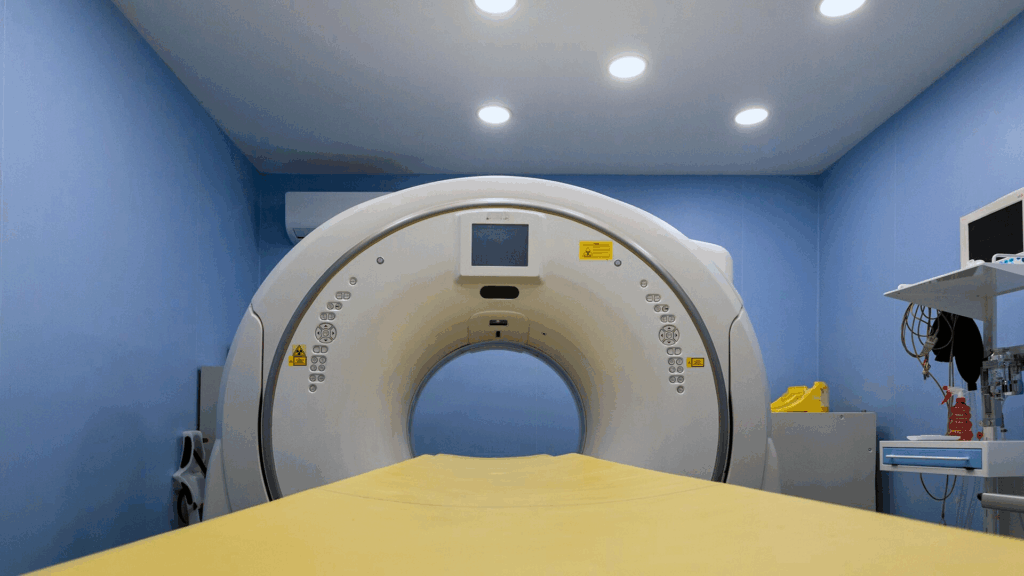 Una macchina per la TAC in una sala medica della Clinica Veterinaria Zarcone, illuminata a giorno e con pareti blu, vista dal letto del paziente che guarda verso l'apertura circolare dello scanner con le attrezzature mediche visibili sul lato destro.