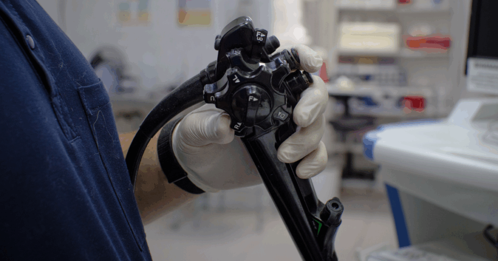 Una persona che indossa un guanto tiene in mano un dispositivo endoscopico veterinario in un ambiente clinico, con attrezzature e forniture mediche sfocate visibili sullo sfondo.